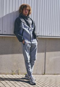 Gray and black windbreaker with geometric design, paired with matching pants; both made of lightweight fabric with zippers and pockets.