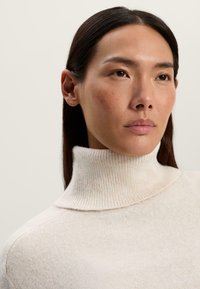 Maglione a collo alto a coste crema con una texture morbida e leggere variazioni di colore. Presenta una vestibilità aderente e spalle senza cuciture.