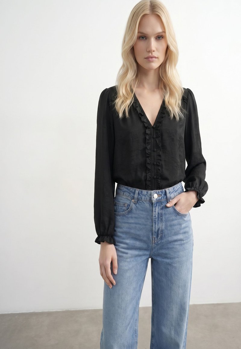 Jeune femme aux longs cheveux blonds portant un chemisier noir à volants et un jean bleu clair taille haute, debout devant un fond uni.