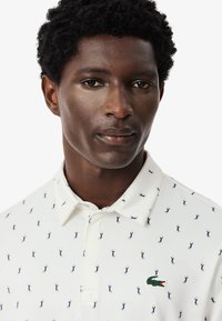 Valkoinen polo-paita, jossa on tummansiniset siluetit kuosina, vasemmassa rinnassa vihreä krokotiililogo ja perinteinen kauluksen muotoilu.