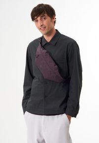 Lila Umhängetasche aus Nylon mit glatter Textur, ausgestattet mit zwei runden Akzenten und einem verstellbaren Schulterriemen. Über einem dunklen Shirt getragen.