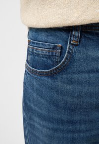 Jean en denim avec un lavage bleu foncé, comprenant une poche avant, des surpiqûres orange contrastantes et une texture lisse.
