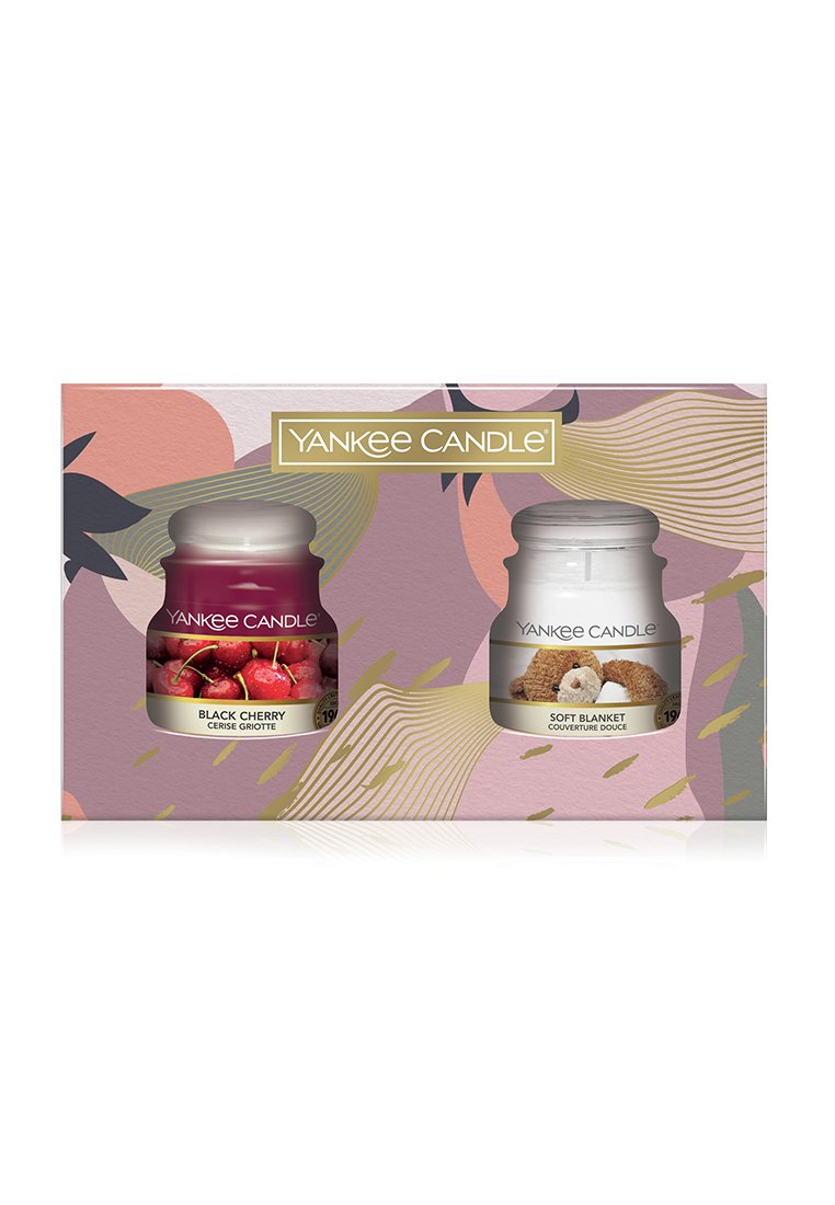 Juego de regalo de Yankee Candle que incluye dos tarros de vidrio: cereza negra (rojo oscuro) y manta suave (blanco) sobre un fondo estampado en rosa y dorado.