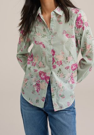 Vrouw draagt een lichtgroen overhemd met knopen met roze en rode bloemenpatronen, gecombineerd met een blauwe spijkerbroek tegen een neutrale achtergrond.