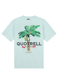 Camiseta de manga corta verde menta con un gráfico de palmera y flamencos, texto "QUOTRELL RESORT" y detalles en amarillo brillante.