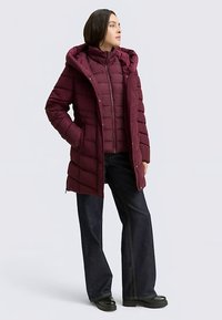 Bourgondische donzen jas met capuchon, voorzien van verticale stiknaden en een ritssluiting, gedragen over een vest in een vergelijkbare kleur, gecombineerd met donkere spijkerbroeken.