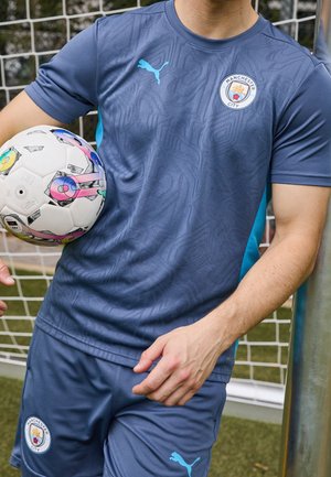 Person wearing blue Manchester City soccer kit holding a colorful soccer ball against a goalpost on a field.