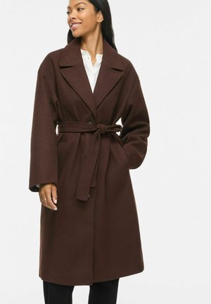 Femme portant un manteau ceinturé marron foncé mi-longueur par-dessus une chemise blanche, debout, une main dans la poche, souriante et regardant sur le côté.