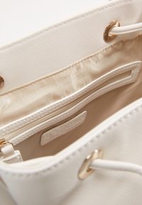 Vit tygväska inuti med en slät beige foder, en ficka med dragkedja och ett öppet fack med en texturerad etikett.