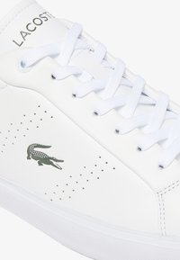 Sneaker bianca in pelle con dettagli traforati, lacci bianchi piatti e logo del coccodrillo Lacoste verde sul lato esterno. Suola in gomma.