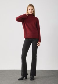 Burgundi színű garbós pulóvert, fekete, bővülő farmernadrágot és fekete magassarkú csizmát viselő nő, aki az egyik kezét a nyaka közelében tartja.