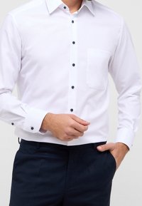 Camisa de dress de manga larga blanca con botones azul oscuro, un bolsillo en el pecho a la izquierda y puños abotonados, combinada con pantalones oscuros.
