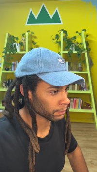 Τζιν καπέλο μπέιζμπολ με ένα αυτοκόλλητο στο μπροστινό μέρος, φορεμένο από άτομο με μακριές dreadlocks. Φωτεινός κίτρινος τοίχος και ράφια με φυτά στο φόντο.