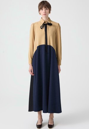 Moteris stovi ilgoje suknelėje, kurios viršutinė dalis šviesiai ruda, apatinė dalis - tamsiai mėlyna, juodos spalvos šoninis panele, juodas kaklaraištis prie apykaklės ir juodi aštrūs bateliai.