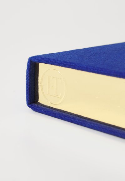 Blue fabric-covered book with gold-edged pages and embossed logo on the spine. Textured cover and neatly stitched edges.