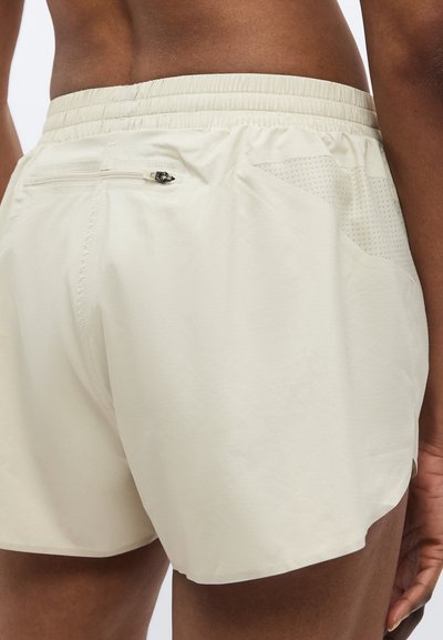 Μπεζ αθλητικό σορτς με ελαστική μέση, διαθέτει πίσω τσέπη με φερμουάρ και δίχτυ κοντά στις τσέπες για καλύτερη αναπνοή.