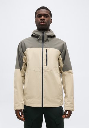 Bărbat purtând o jachetă bej și verde măsliniu cu glugă, cu fermoar frontal și buzunar cu fermoar pe piept, stând în fața unui fundal simplu.