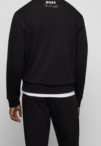 Sudadera negra con bajo y puños acanalados, con un logotipo dorado en la parte posterior del cuello. Se completa el conjunto con pantalones negros.