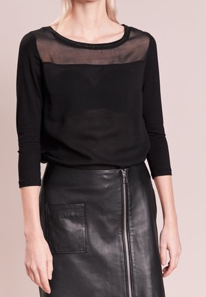 Femme portant un haut noir transparent à manches trois-quarts et une jupe en cuir noire avec fermeture éclair devant et poche plaquée.