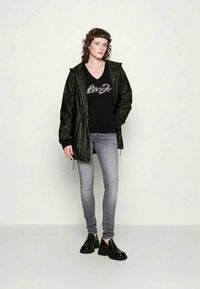 Fekete, mintás, könnyű dzseki, húzózsinórral, egy fekete V-nyakú póló fölött, amely fényes felirattal rendelkezik. Szürke farmer és fekete platform cipő.