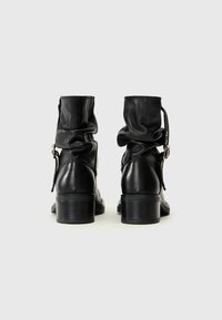 Bottes noires en cuir à la cheville avec un design plissé, détail de boucle et talon empilé. Texture lisse et bout arrondi.