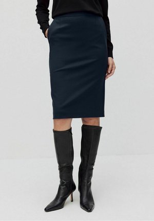 Mujer con falda lápiz azul marino oscuro con bolsillos laterales y botas negras de cuero hasta la rodilla con tacón, sobre un fondo liso.
