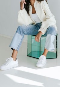 Białe tenisówki z teksturowanymi cholewkami; niska sylwetka. Noszone z jasnoniebieskimi, skróconymi dżinsami oraz kremowym swetrowym żakietem. Przezroczysty, geometryczny szklany stół.