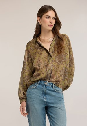 Vrouw die een groen blouse met paisley-patroon draagt, gecombineerd met een blauwe spijkerbroek, staat met één hand langs haar zij en kijkt naar rechts.