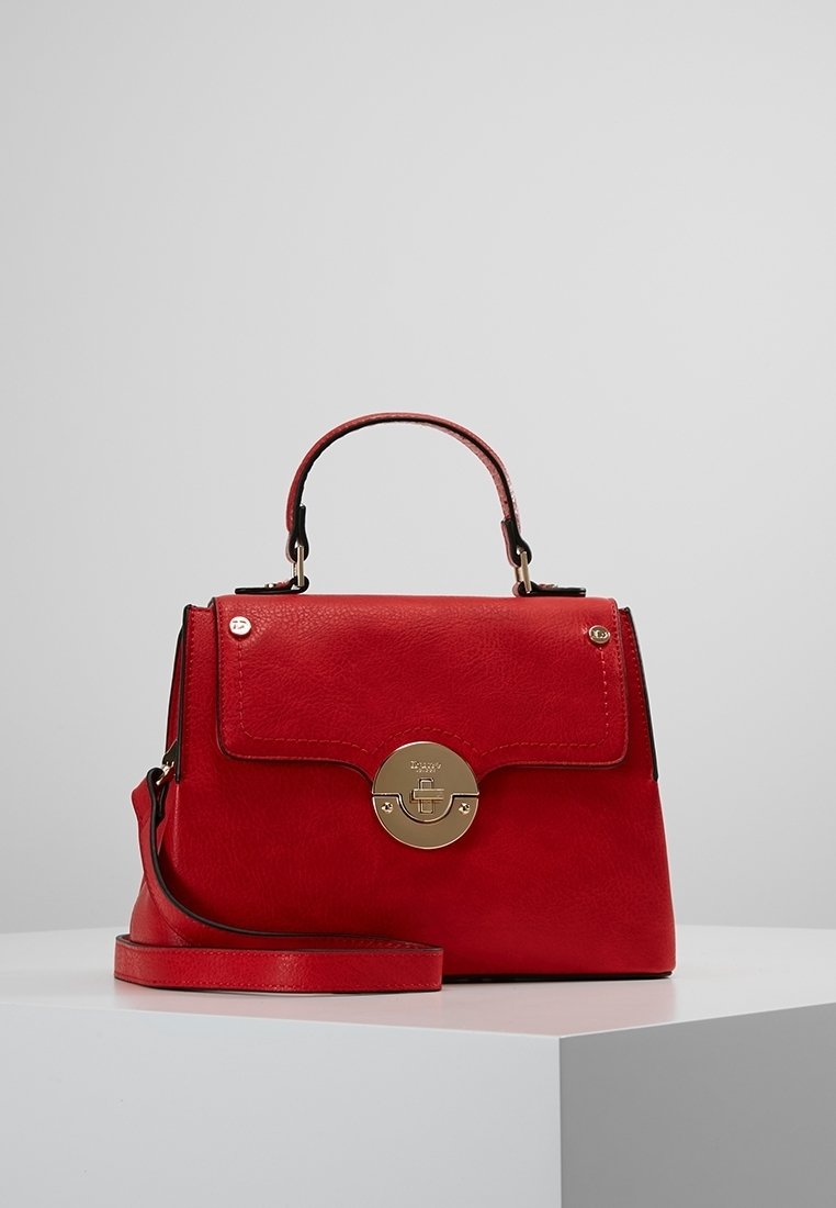 Sac à main en cuir rouge avec poignée supérieure, bandoulière amovible et fermoir rond doré sur surface blanche sur fond gris.
