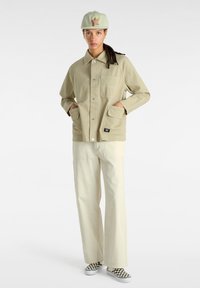 Chaqueta de color beige claro con cuello, bolsillos frontales y cierres a presión, combinada con pantalones color crema y zapatos a rayas en blanco y negro.