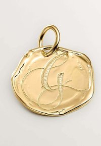 Colgante de oro con forma redonda irregular y texturizada que presenta una letra "G" en cursiva grabada en el centro. Incluye un lazo circular para su sujeción.