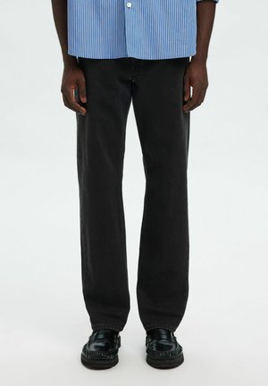 Persona che indossa jeans neri, scarpe di pelle nere e una camicia a righe blu e bianco con le maniche arrotolate.