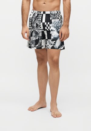 Homme portant un short noir et blanc à motifs représentant des visages de sculptures classiques et des dessins géométriques, debout pieds nus sur un fond uni.