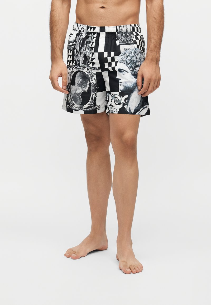 Homme portant un short noir et blanc à motifs représentant des visages de sculptures classiques et des dessins géométriques, debout pieds nus sur un fond uni.