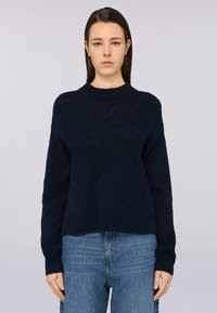Granatowy, teksturowany sweter z okrągłym dekoltem i ściągaczami na rękawach, w zestawie z jasnoniebieskimi dżinsami. Materiał wydaje się miękki i gruby.