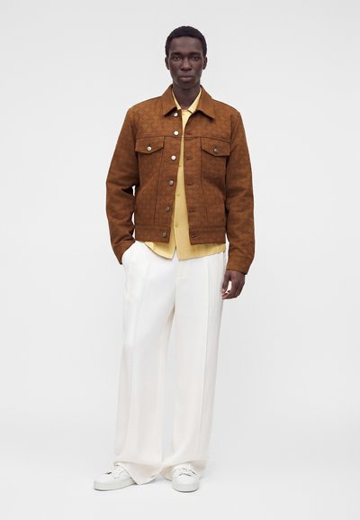 Homme portant une veste marron à motifs, une chemise beige, un pantalon blanc à jambes larges et des baskets blanches, debout avec une main dans la poche.