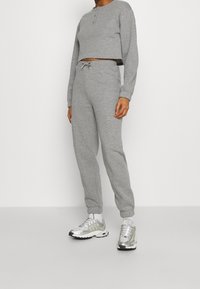 Felpa grigia corta con tre bottoni al collo, abbinata a pantaloni della tuta grigi con vita e polsini elasticizzati. Scarpa da ginnastica argentata con dettagli riflettenti.