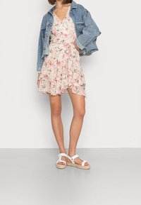 Robe à fleurs rose clair et crème avec ourlet à volants, associée à une veste courte en jean. La tenue est complétée par des sandales blanches à brides.