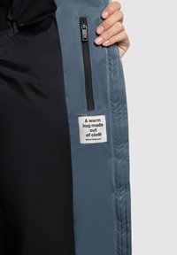 Pehme voodriga jakk, mille sinihall väline ja must sisemine vooder, varustatud lukuga ja kangasildiga, millel on kiri "Soa embrüüs, mis on valmistatud kangast."