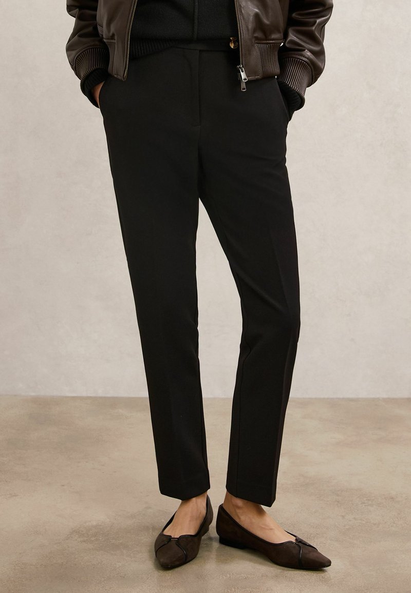 Zwarte, op maat gemaakte broek van zachte stof, met een rechte pasvorm en taille op middelhoog niveau. Gecombineerd met donkerbruine suede ballerina's.