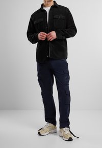 Chemise noire en velours côtelé avec boutons et deux poches poitrine, associée à un pantalon cargo bleu marine avec poches latérales, et des chaussures de sport beiges.