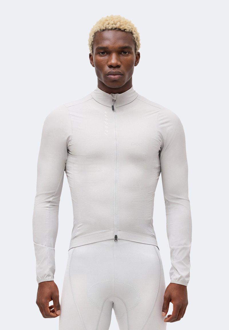 Homme aux cheveux blonds bouclés portant une veste de cyclisme près du corps à manches longues gris clair et un cuissard rembourré assorti.