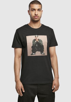 Mann trägt ein schwarzes T-Shirt mit einem aufgedruckten Foto einer Person, die in dunkler Kleidung und Turnschuhen hockt und das Gesicht in den Händen ruhen lässt.