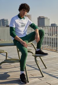 Bílozelená košile Lacoste s logem, zelené joggingové kalhoty a černé tenisky. Venku, na dřevěné terase s kovovými židlemi.