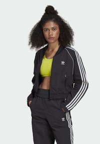 Giacca bomber nera a taglio corto con strisce bianche lungo le maniche, tasche con zip e colletto a coste. Indossata sopra una maglietta gialla brillante.