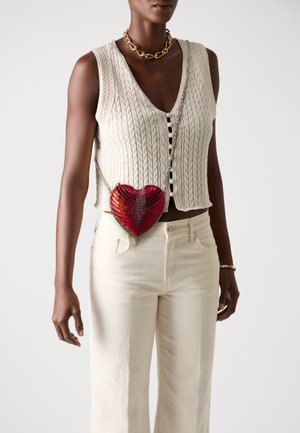 Canottiera in maglia color crema con scollo a V profondo, abbinata a jeans a gamba dritta di colore chiaro. Una borsa a forma di cuore rossa è indossata a tracolla con una catena.