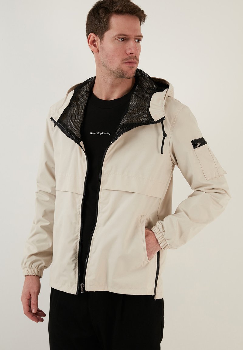 Lichtbeige waterdichte jas met een capuchon, ritsluiting, elastische manchetten, twee voorzakken en een mesh voering. Voorzien van een met een rits afsluitbare mouwzak.
