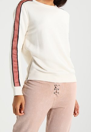 Vrouw draagt een wit shirt met lange mouwen met roze en zwarte gestreepte tekst op de mouwen en lichtroze broek met vetersluiting aan de voorkant.