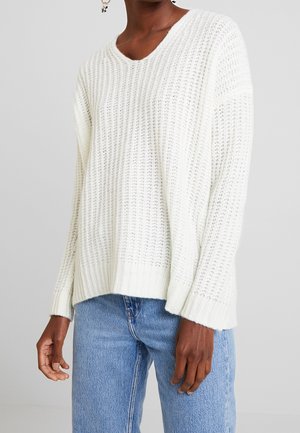 Person iført en løs, hvid striktrøje og lyseblå jeans, stående mod en ensfarvet, lys grå baggrund.