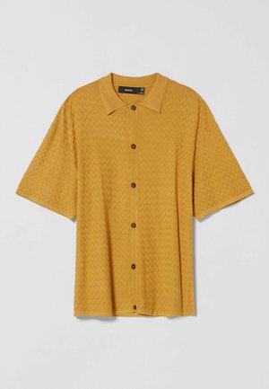 Chemise à manches courtes jaune moutarde avec un motif zigzag subtil et un col classique, présentée sur un fond uni.